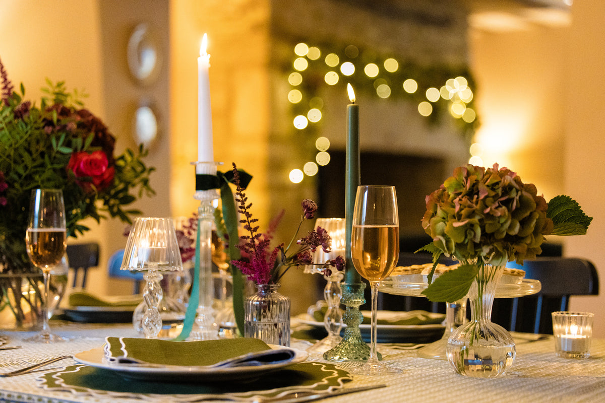 Dining table set with tall glass candlesticks and autumnal flowers in a warm, cozy setting complete with forest fern green table linen.