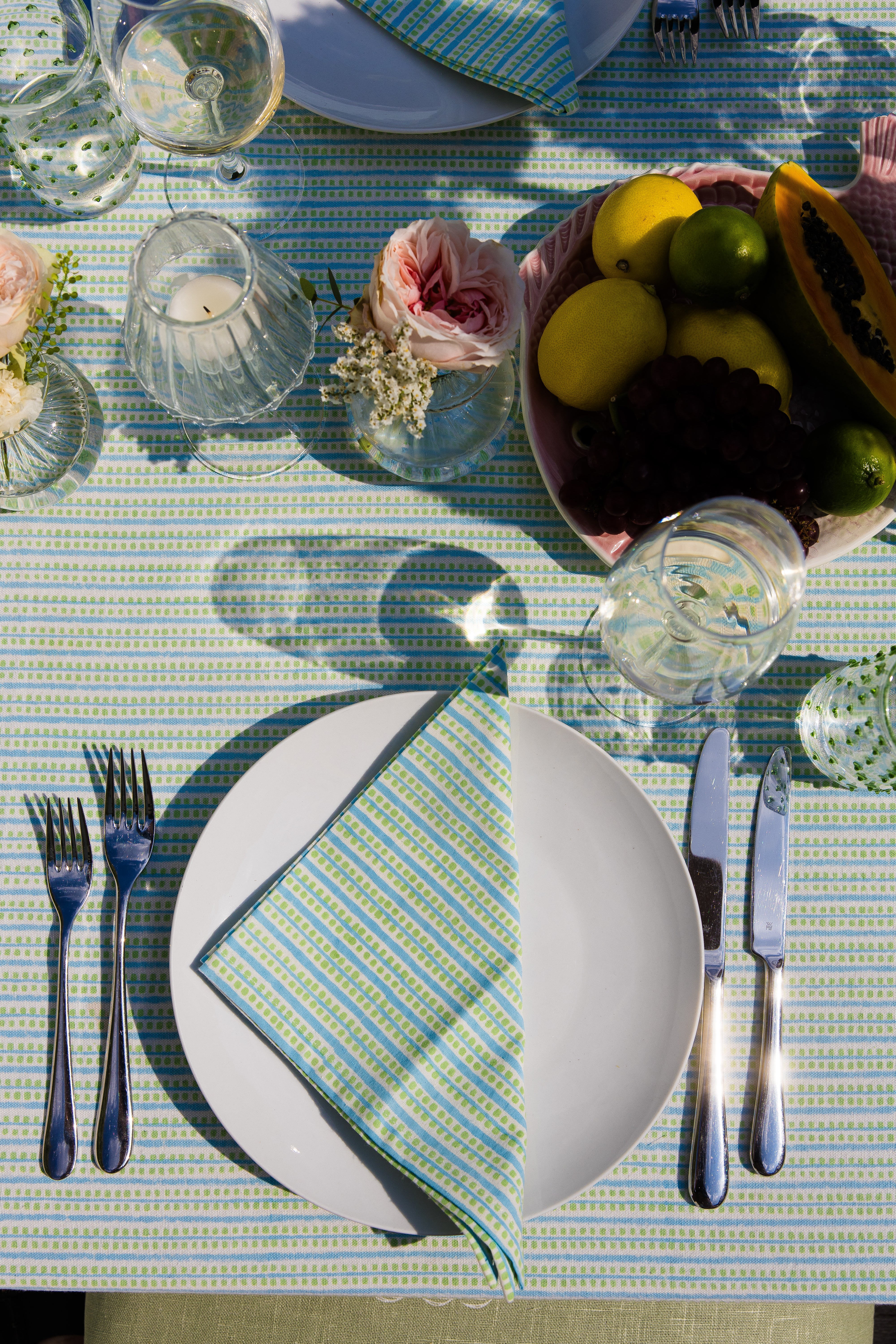 Spring table setting with plates, cutlery, and a bowl of fruit on a striped dotted green and turquoise tablecloth.