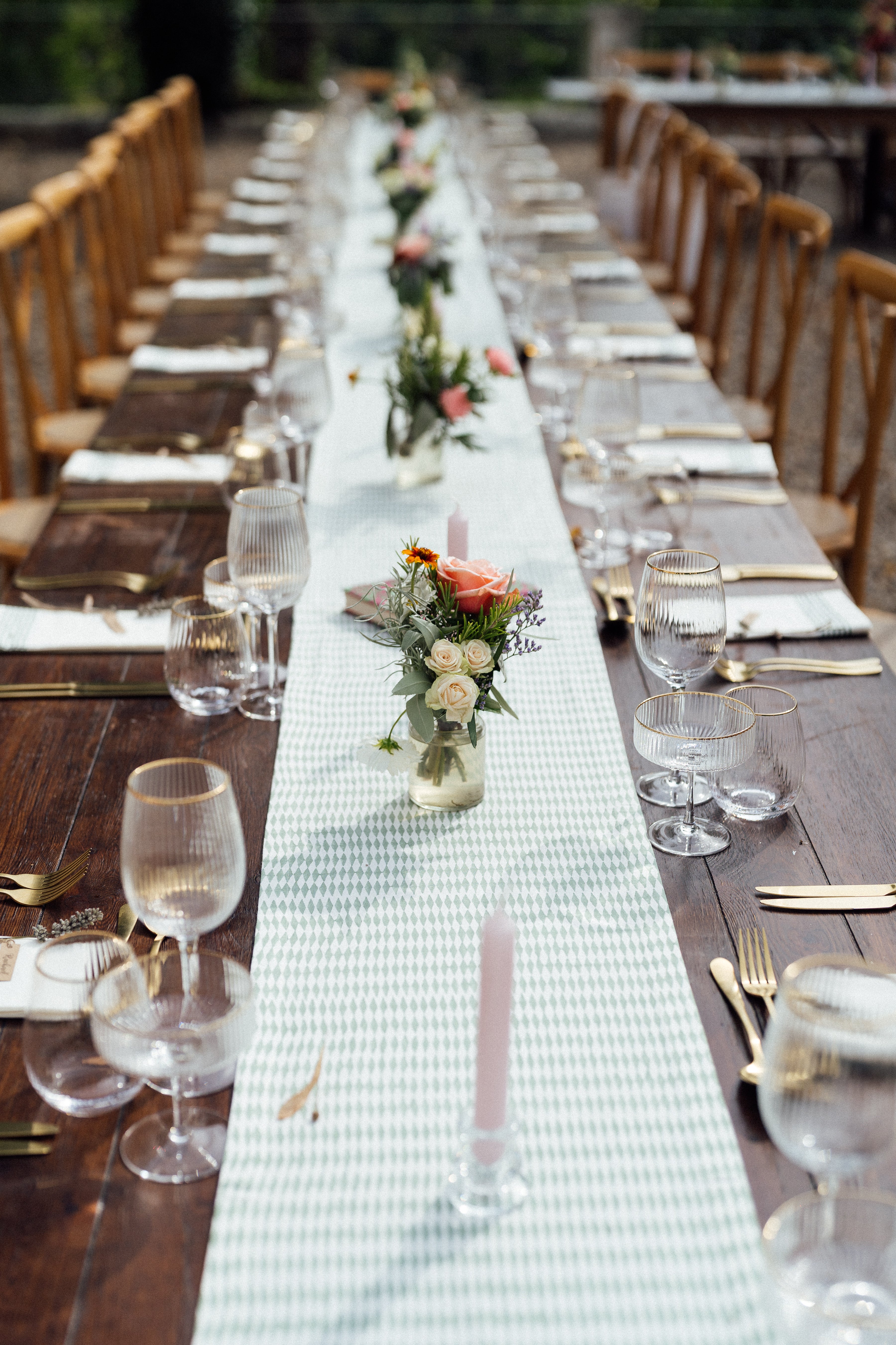 Long table set for a wedding meal with place settings, glasses, and floral centerpieces with a colourful cotton runner and elegant glassware bud vases.