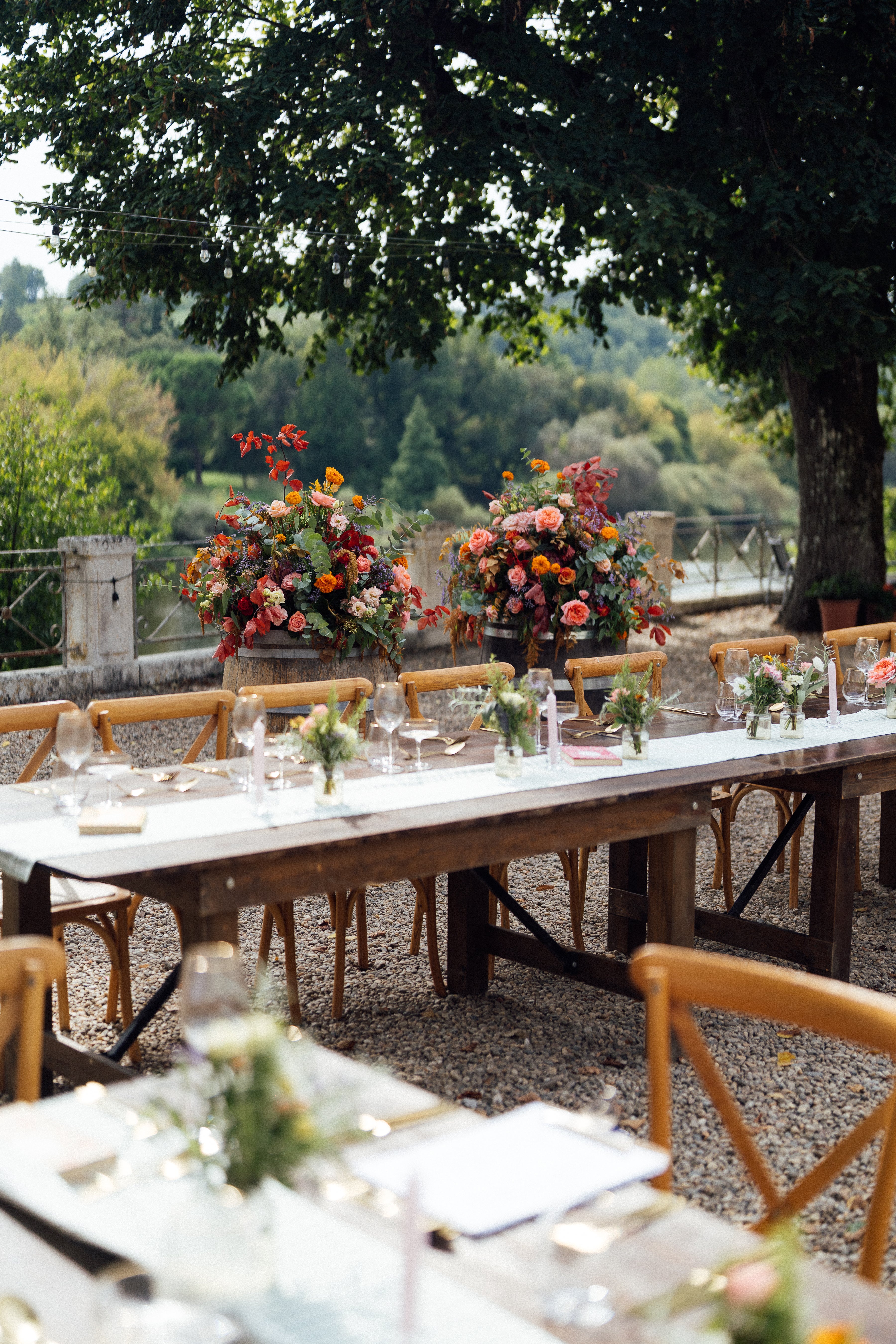 Outdoor setting with a long wooden table set for a wedding, surrounded by chairs and floral arrangements with beautiful glasses.