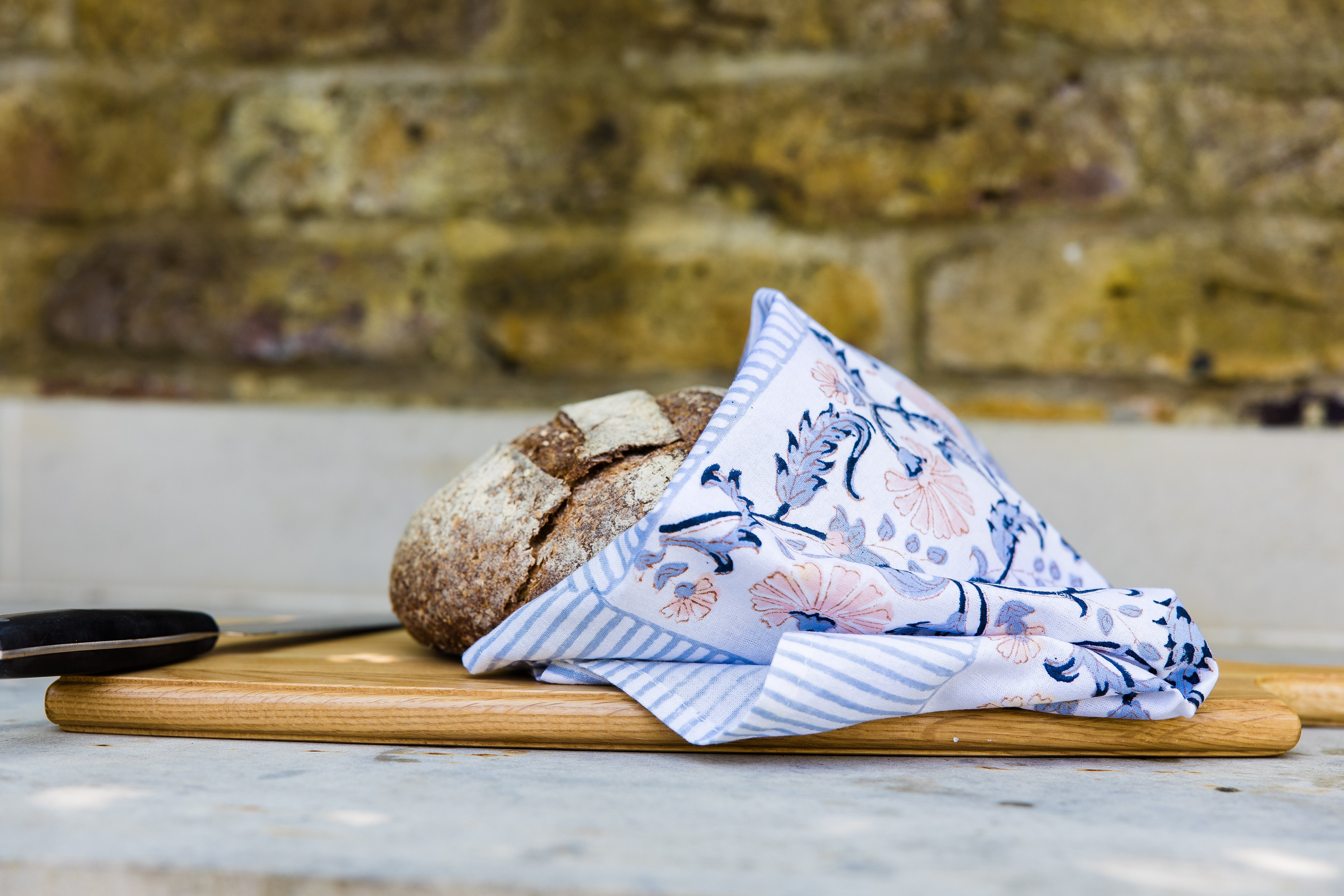 Bread wrapped in a floral kitchen towel on a wooden cutting board with a brick wall background