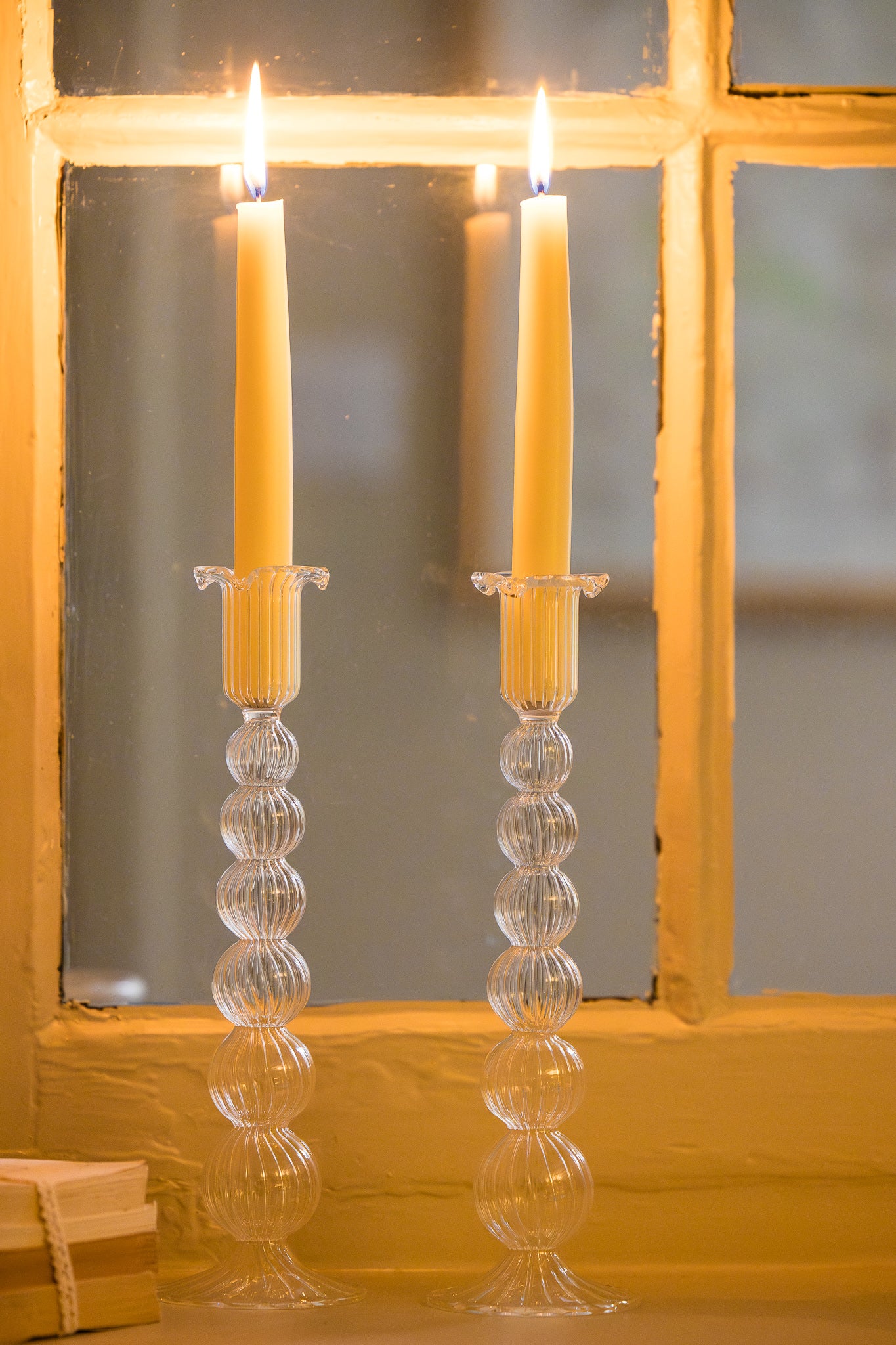 Two tall, thin glass bubble candle holders with lit candles in front of a  window.